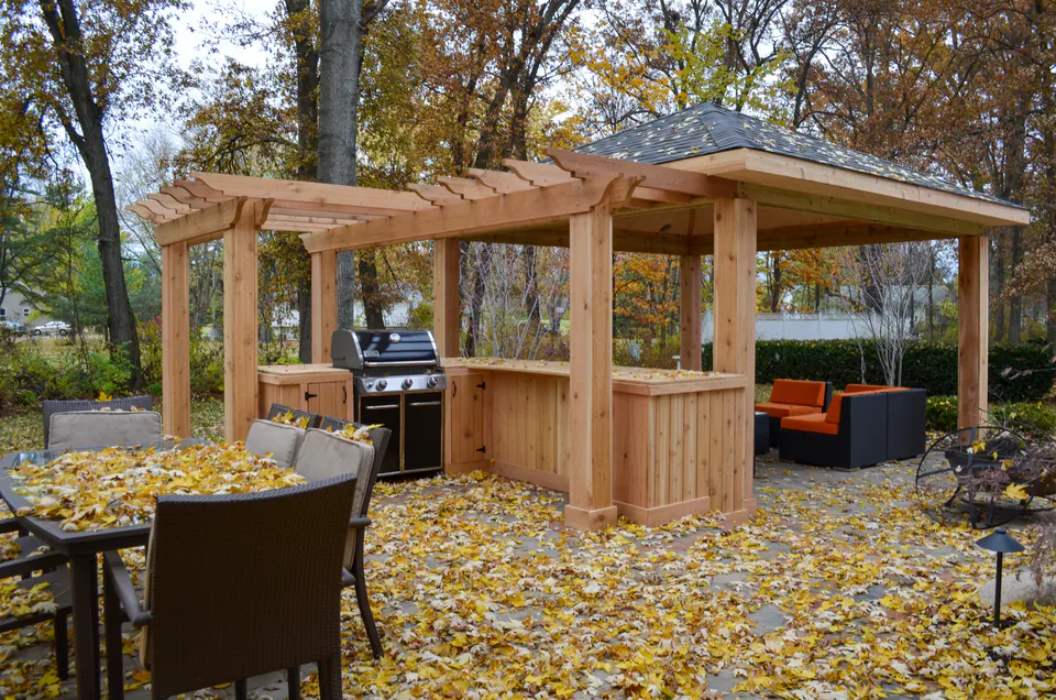 Outdoor kitchen and pergola-style pavilion with grill, wood counter, paver patio, dining chair, and fall leaves.